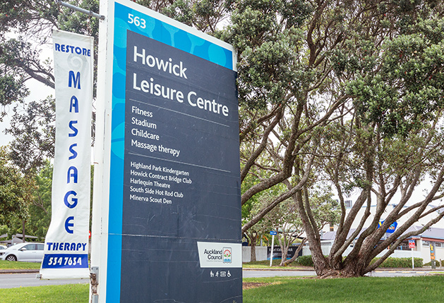 Tall blue and grey Howick Leisure Centre sign at 563 with a wheelchair symbol at the base. A white vertical massage therapy banner hangs to the left, beside large leafy trees and green grass.