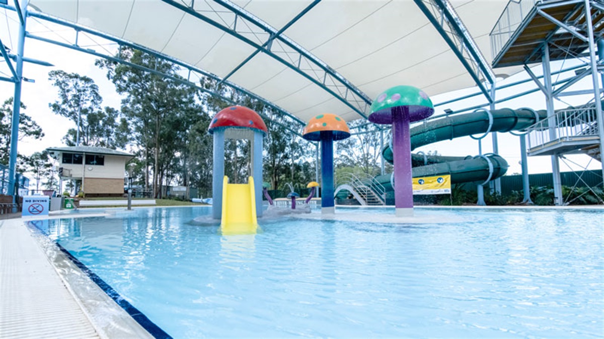 Tiled pool edge with 'No Diving' sign. Shallow pool features colourful mushroom fountains and a yellow slide. Metal stairs with handrails lead to a green water slide under a white shade canopy.
