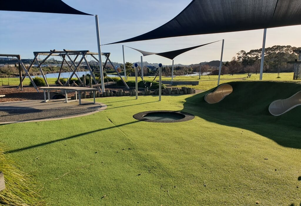 Level green synthetic turf with a flush in-ground trampoline. Curved timber bench on a concrete path near wooden swing frames. Two tunnel slides in a grassy mound under black shade sails.