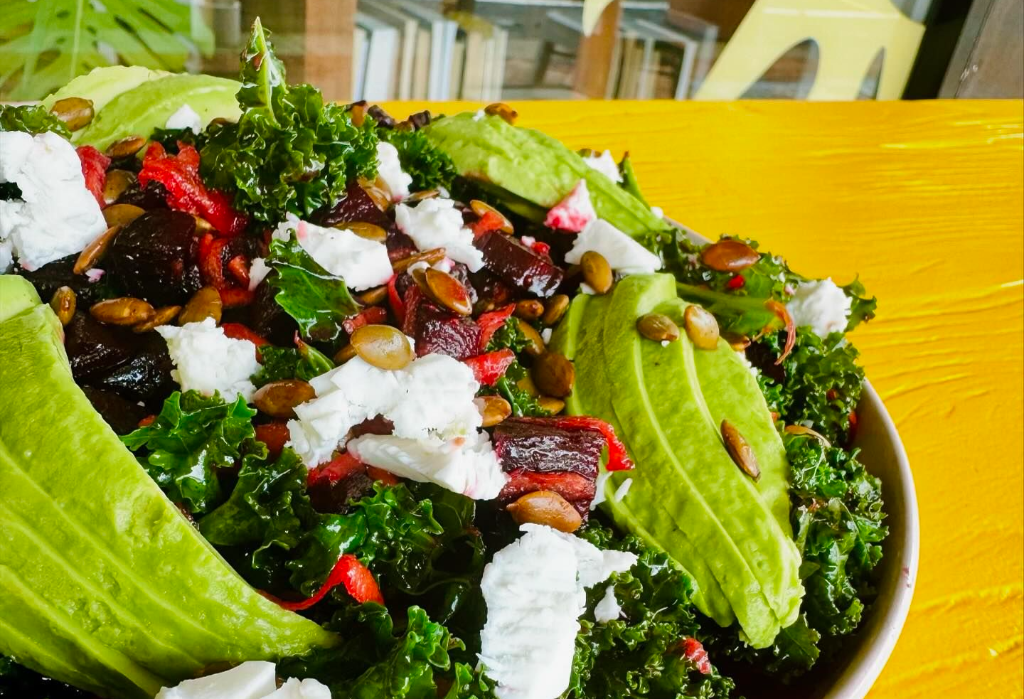 Vibrant salad with green kale, fanned avocado slices, dark beetroot chunks, white cheese, and toasted seeds in a white bowl on a bright yellow table.