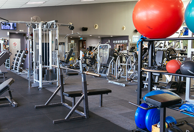 Level gym floor with black rubber mats and grey carpet. Wide, step-free pathways between exercise machines and benches. Bright overhead lighting and clear signage on background doors.