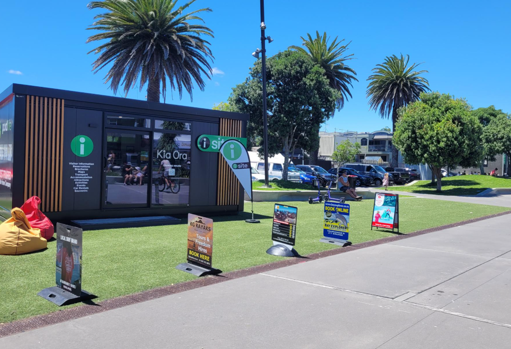 Portable information building with level entry from synthetic turf. Wide concrete path in foreground. A-frame signs and bean bag seating on grass. Green 'i-site' and 'Kia Ora' signage.