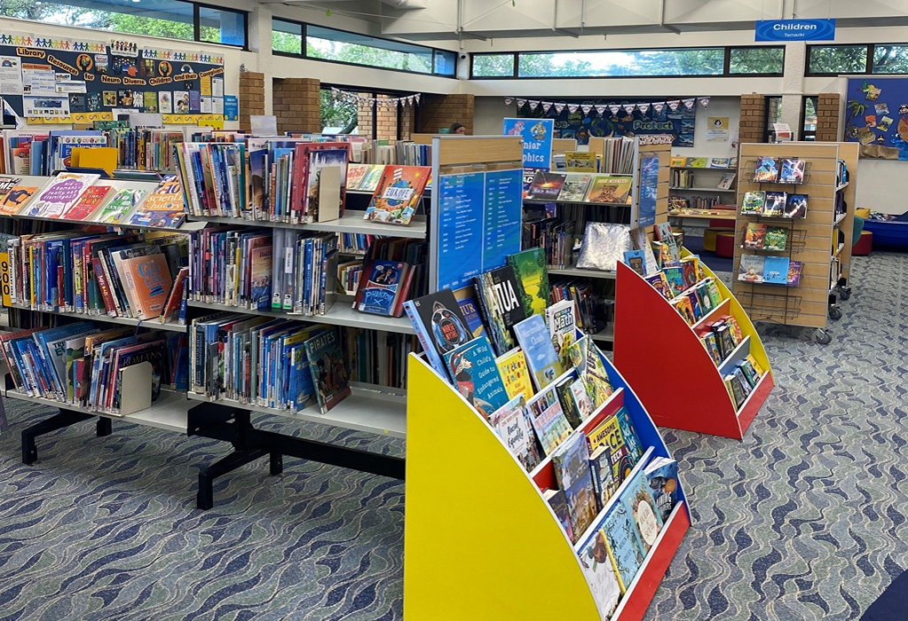 Level blue and grey patterned carpet with wide pathways between low-level metal shelving and colourful slanted display units. Brightly lit room with a blue 'Children' sign overhead.