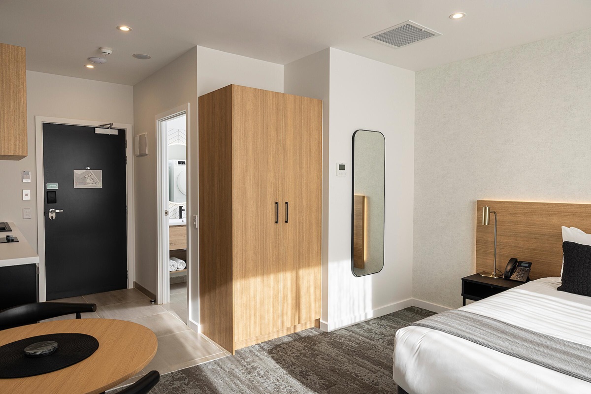 Level tiled floor transitions to grey carpet. Black door has a silver lever handle. Open doorway leads to a tiled bathroom. Clear pathways around the dining table, wardrobe, and bed.