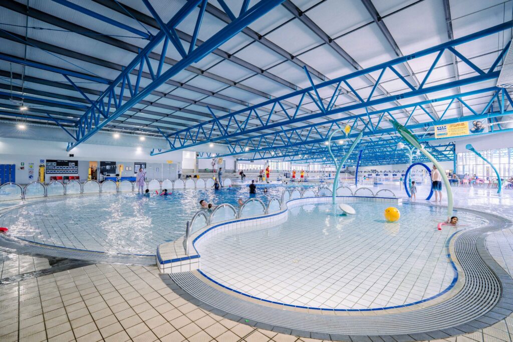Indoor aquatic centre with light grey tiled floors and blue roof trusses. Curved stainless steel handrails line a shallow ramp into a tiled leisure pool with level deck access.