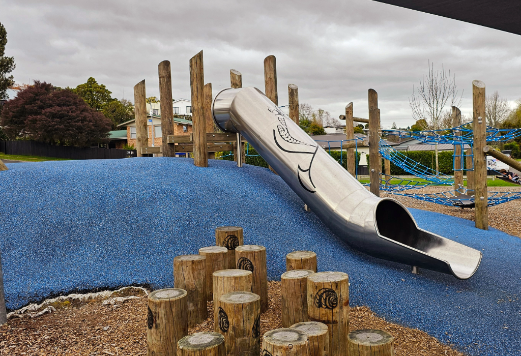 Metal tunnel slide on a blue rubber soft-fall mound. Wood chip ground features carved timber stumps and a blue rope climbing structure supported by tall timber poles.