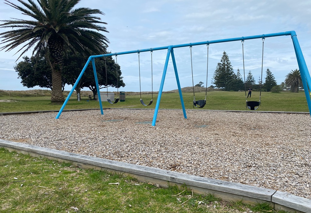 Blue metal swing set with two belt and two bucket seats over woodchips, enclosed by a low timber border. Surrounding ground is level grass with no visible paved pathways.