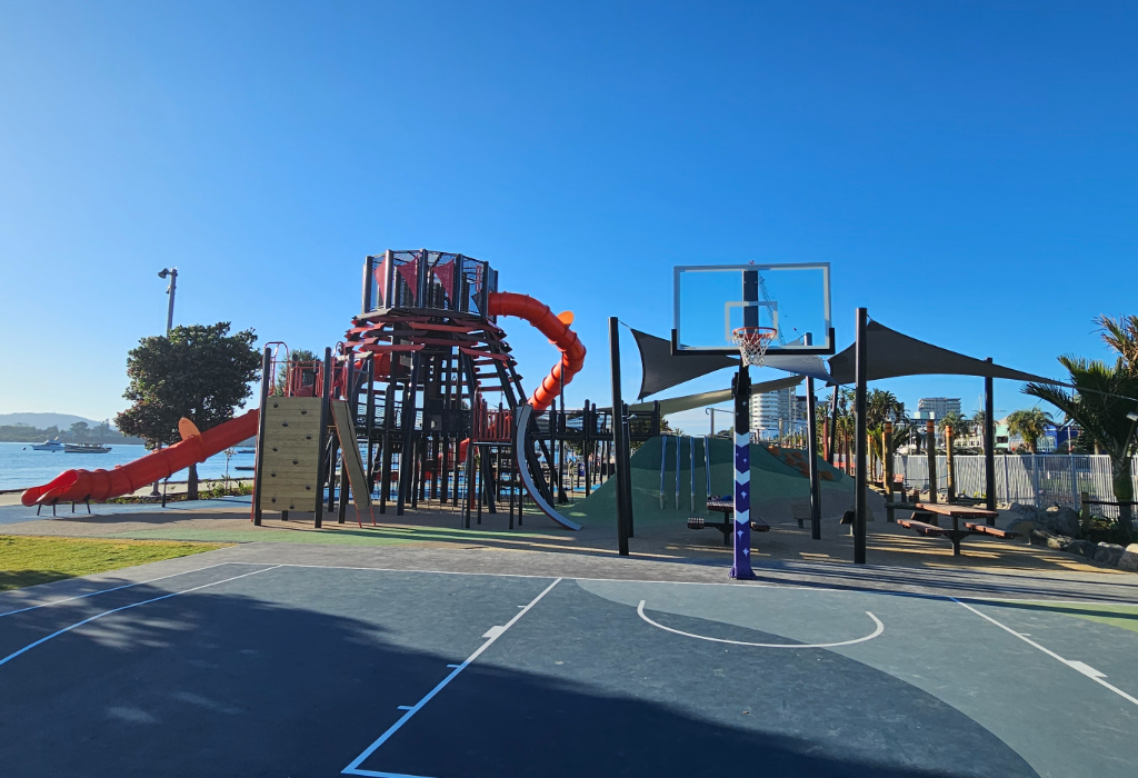 Wide, level blue and grey paved basketball court connects to an open playground with red slides. Timber picnic tables are on flat ground. Unobstructed, level pathways throughout the outdoor space.