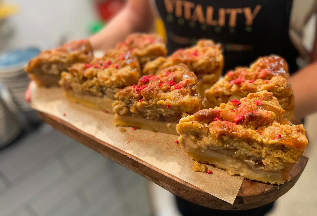 Golden-brown fruit crumble slices topped with red sprinkles on a wooden board. In the background is a level grey tiled floor and stacked plates in a well-lit indoor area.