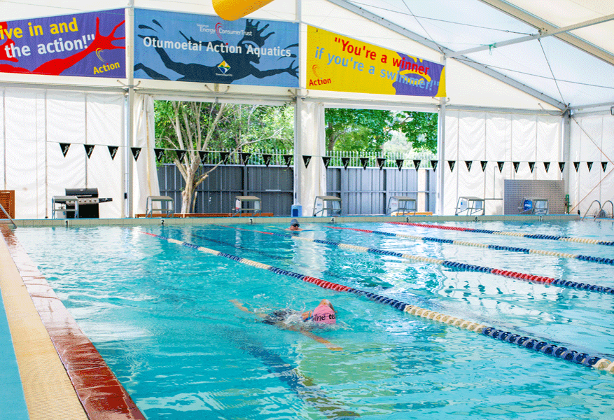 Indoor pool with level access, textured beige non-slip tiles and red edge. Blue and red lane ropes divide the water. High white canopy roof and bright wall banners above the pool deck.