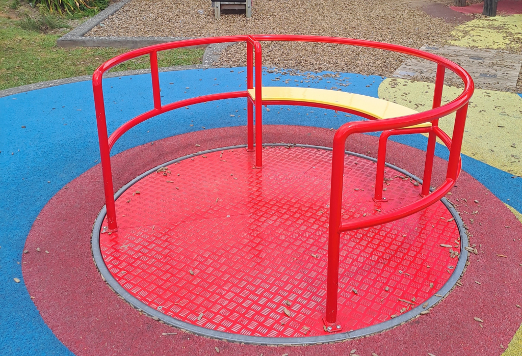 Level-access red carousel on flat multi-coloured rubber flooring. Features a textured metal platform, red perimeter handrails with one wide entry point, and a curved yellow bench seat.
