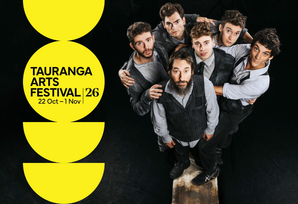 Yellow graphic signage for Tauranga Arts Festival, 22 Oct – 1 Nov. Six men stand on a narrow wooden beam on a dark, level floor surface.