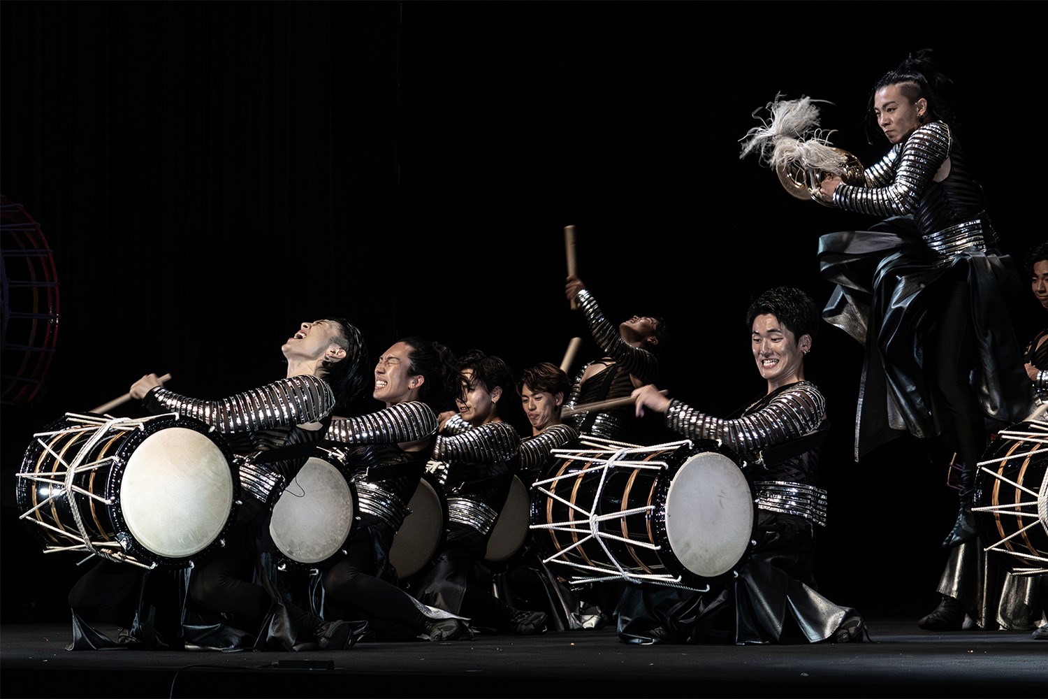 Performers in silver and black striped outfits play large white drums on a level, dark stage. One person jumps mid-air. The surface is flat and matte black with no visible obstructions.