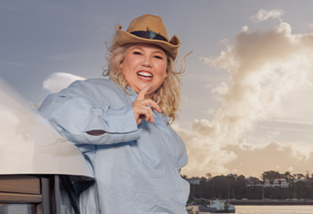 Blonde woman wearing a straw cowboy hat and light blue shirt, leaning against a white vehicle. Harbour water and distant shoreline under a cloudy sky in the background.