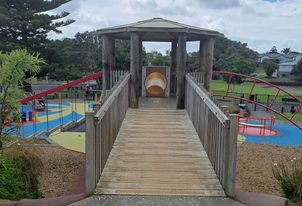 Wide wooden ramp with yellow non-slip strips and high timber handrails leads to a gazebo. Level entry from a path. Blue and yellow rubberised safety surface and bark mulch below.