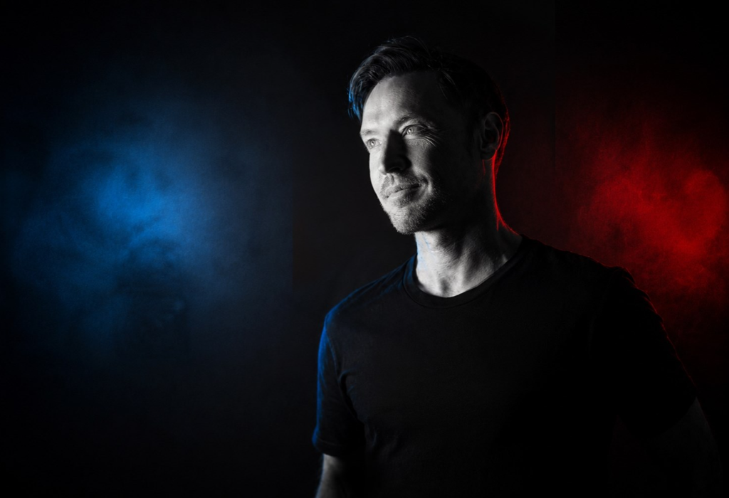 Man with short dark hair and stubble wearing a black t-shirt. Blue and red lighting from the sides highlights his face against a dark, smoky background with misty colour effects.