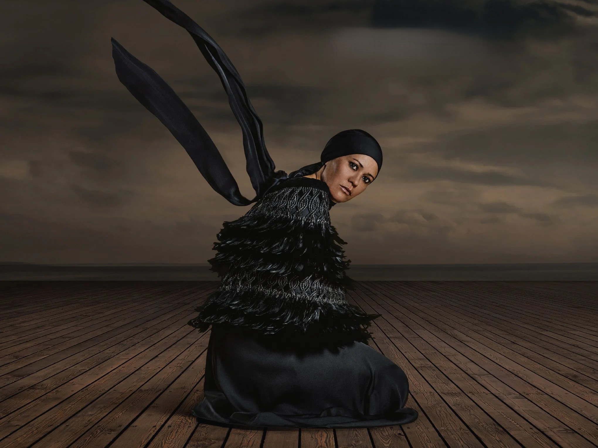 Person wearing a black feathered cape and headscarf kneels on a level wooden plank boardwalk. Long black ribbons flow upwards against a dark, cloudy sky over the sea.