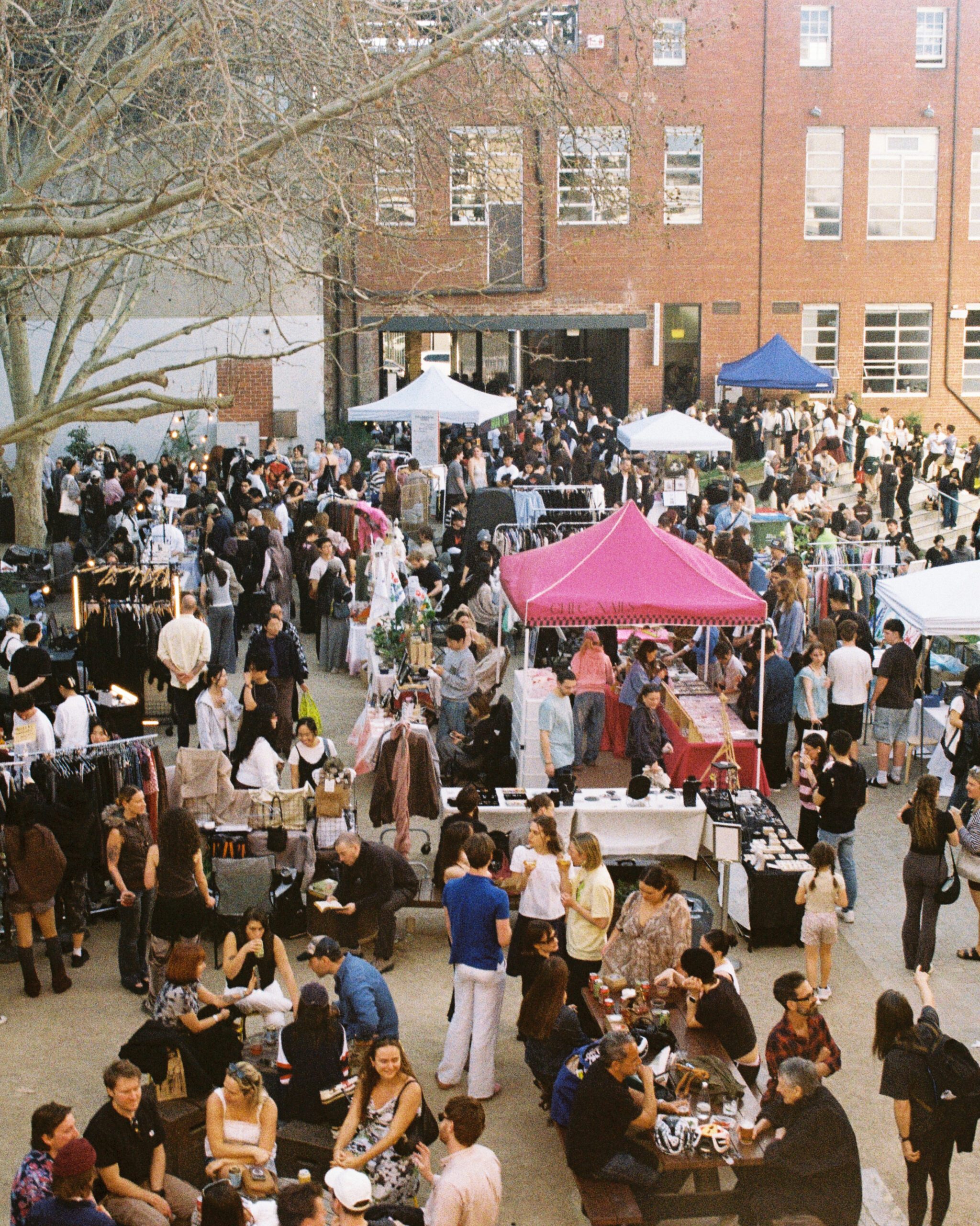 Level paved courtyard with wooden picnic tables. White and pink market stalls sit on flat ground. A wide, step-free building entrance is visible in the background.
