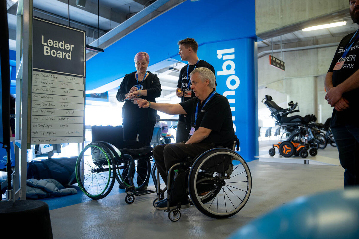 Level polished concrete floor in a bright indoor area. A man in a manual wheelchair reaches toward staff near a white Leader Board sign. A power wheelchair is parked in the spacious background.
