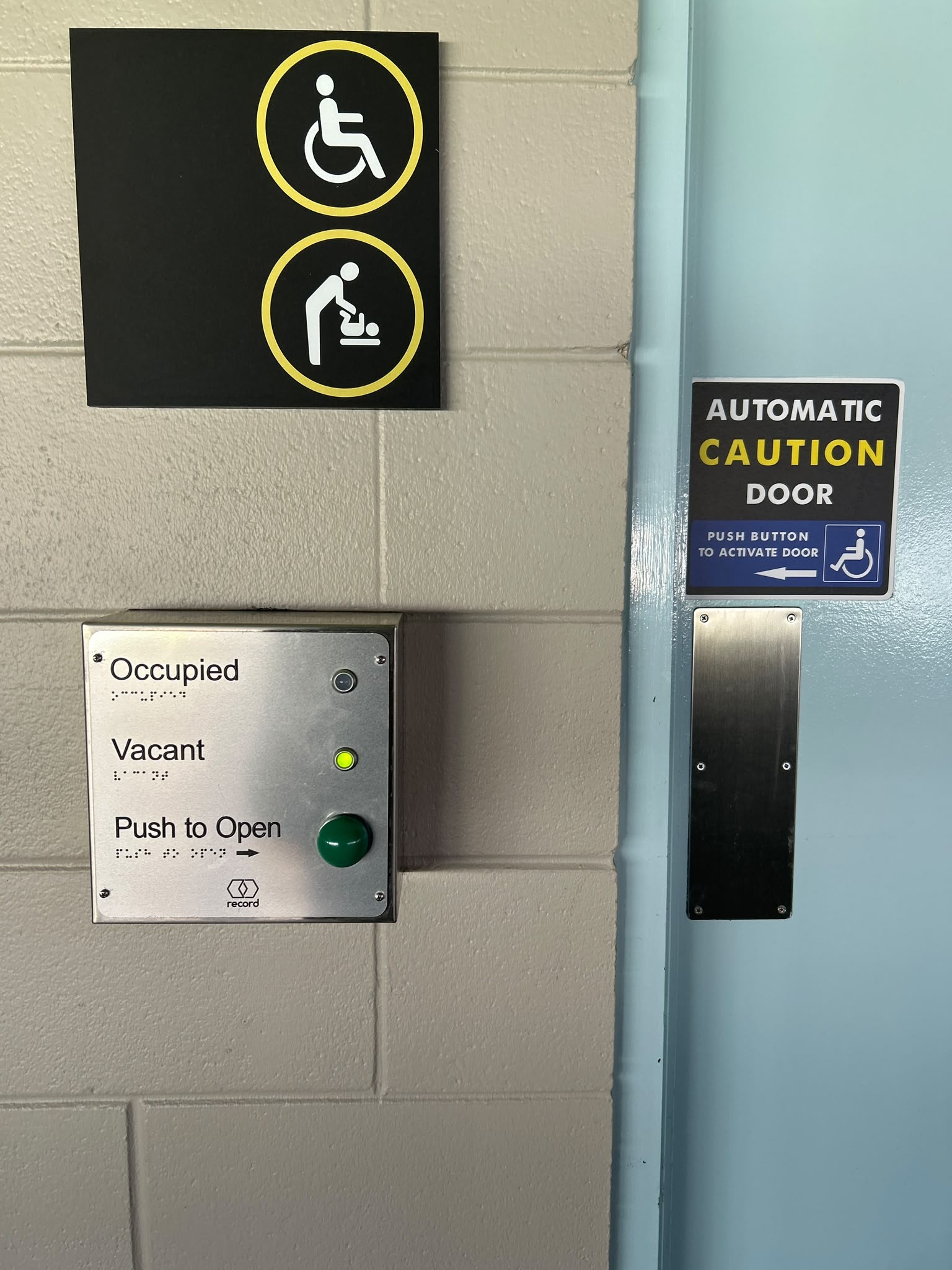 Accessible entrance with light blue automatic door. Metal panel on grey wall features braille, a green ‘Vacant’ light, and a push button below wheelchair and baby change signage.