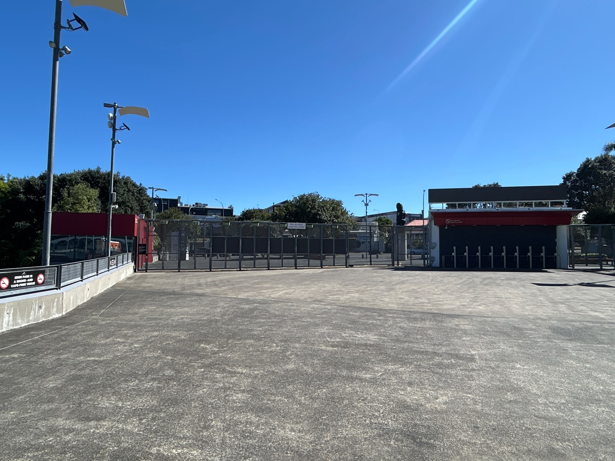 Wide, level concrete area leads to black metal security gates. A red ticketing building with turnstiles stands on the right. A low concrete wall with signage borders the left under a clear sky.