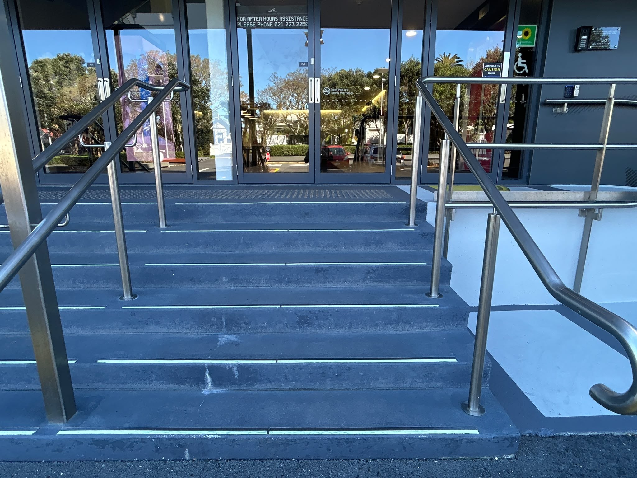 Five blue steps with light-coloured edge strips lead to automatic glass doors. Stainless steel handrails on both sides and along a concrete ramp on the right. Wheelchair symbol on entrance door.