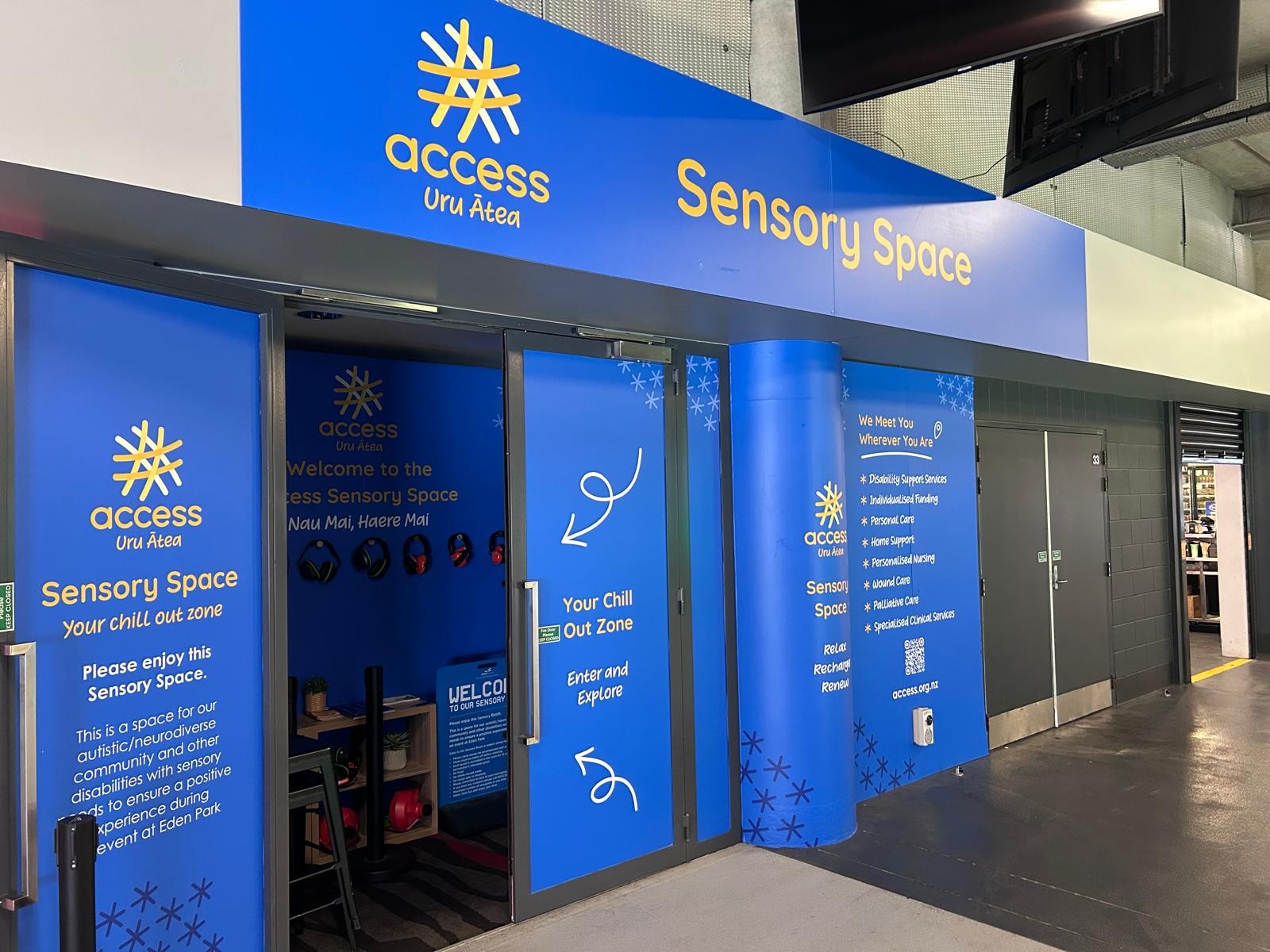 Wide level entrance to a blue Sensory Space with glass doors and yellow signage. Smooth grey flooring transitions to carpet inside. Noise-cancelling headphones hang on the wall at waist height.