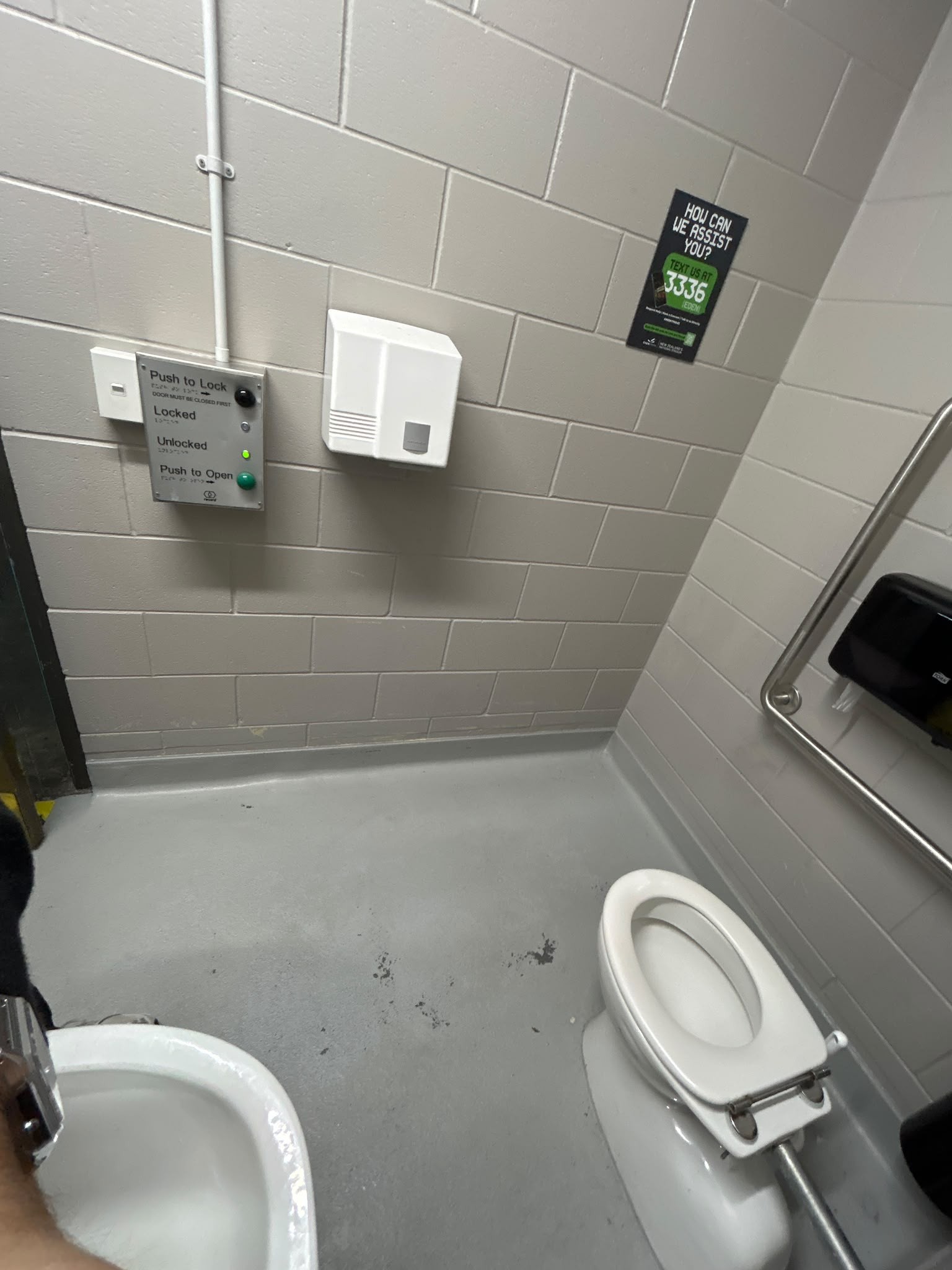 Accessible toilet with grey block walls, concrete floor, and metal grab rails. Electronic door control panel with push buttons, white hand dryer, and black paper towel dispenser.