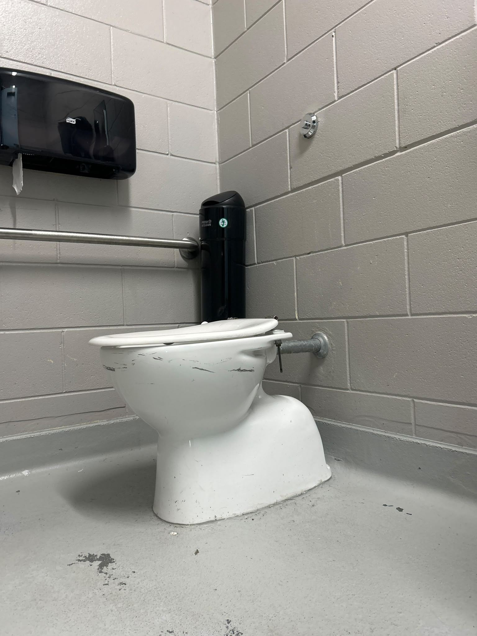 White toilet on level grey floor. Horizontal silver grab bar on left grey brick wall. Small silver flush button on right wall. Black paper towel dispenser and slim black bin nearby.