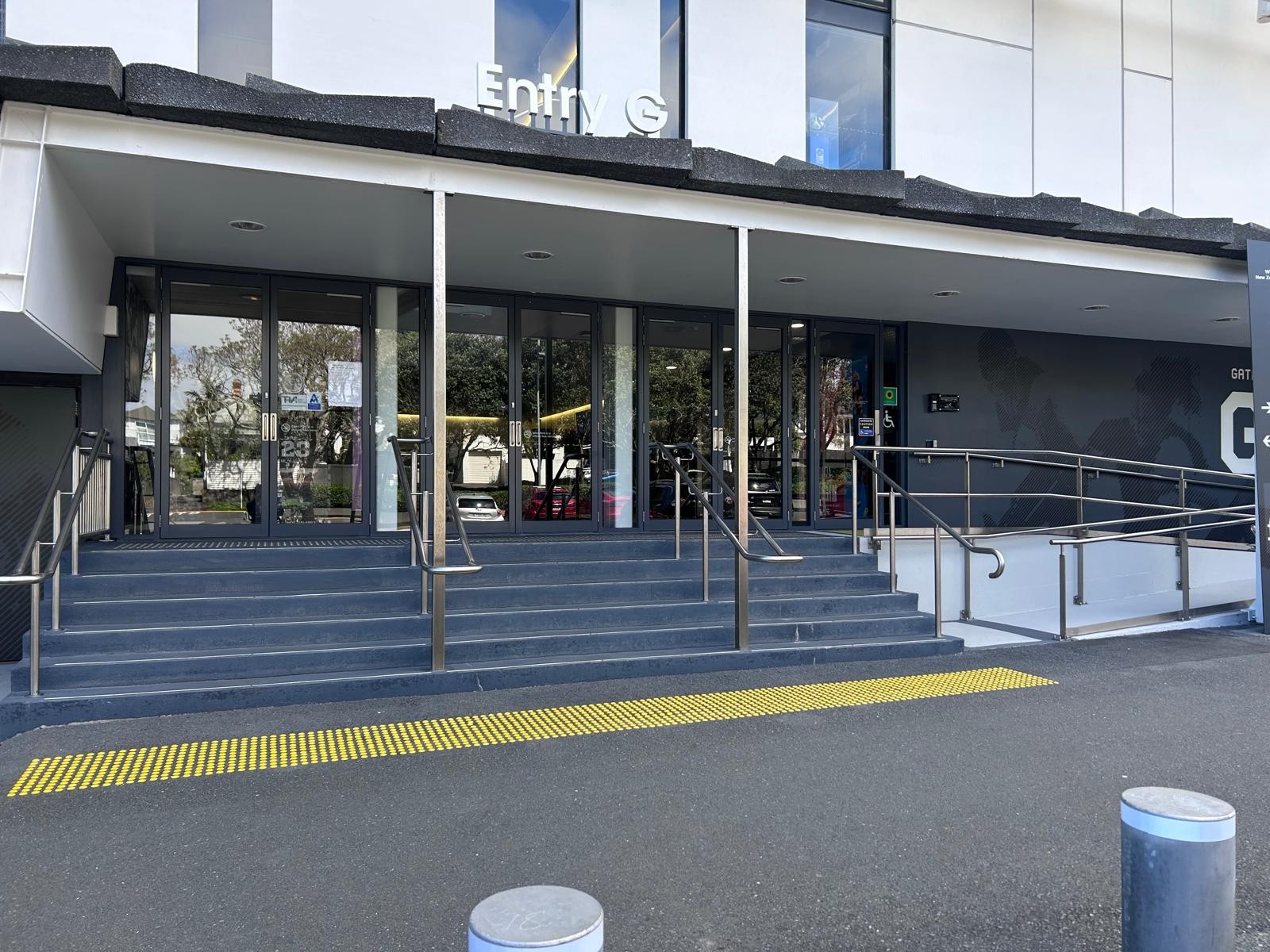 Entry G signage above glass doors. Grey stairs with yellow tactile strip and stainless steel handrails. Concrete ramp with handrails on the right leads to a level automatic door.