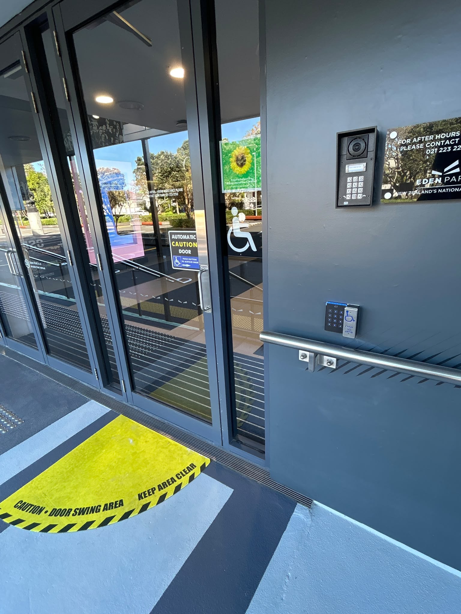 Level glass entrance with automatic doors, tactile ground indicators and yellow 'DOOR SWING AREA' markings. Lowered keypad with wheelchair symbol and metal handrail on the dark grey wall.