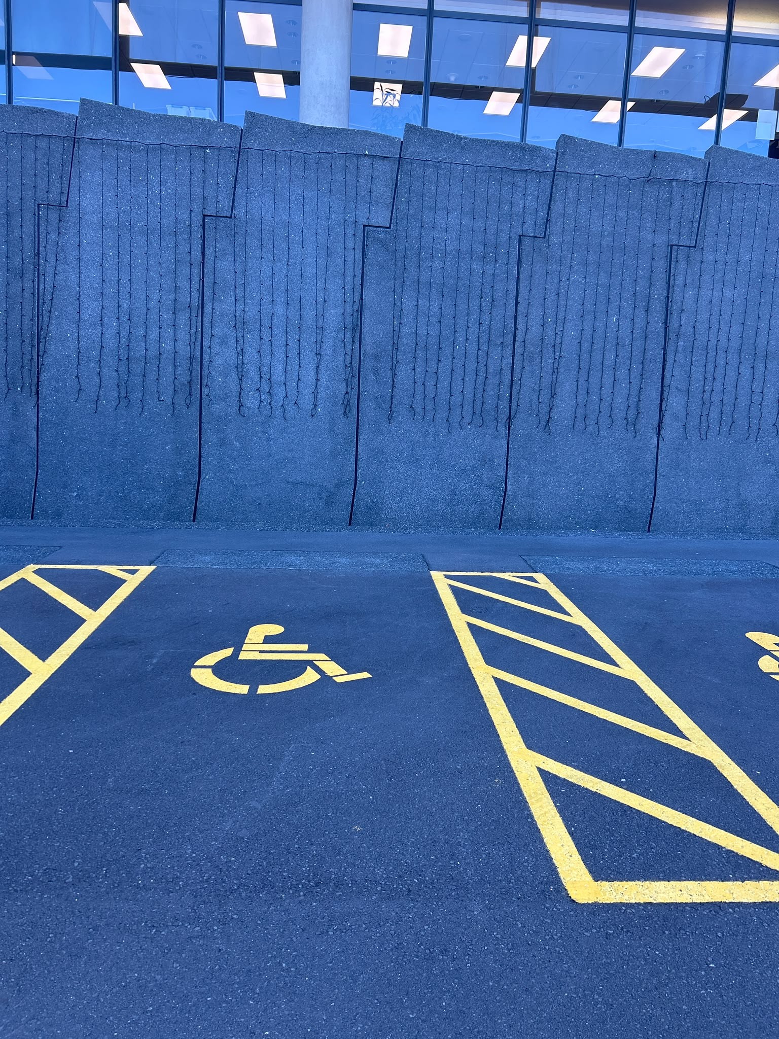Level dark asphalt accessible parking bay with a yellow wheelchair symbol and hatched access aisle. A textured grey concrete wall and building windows are behind the space.