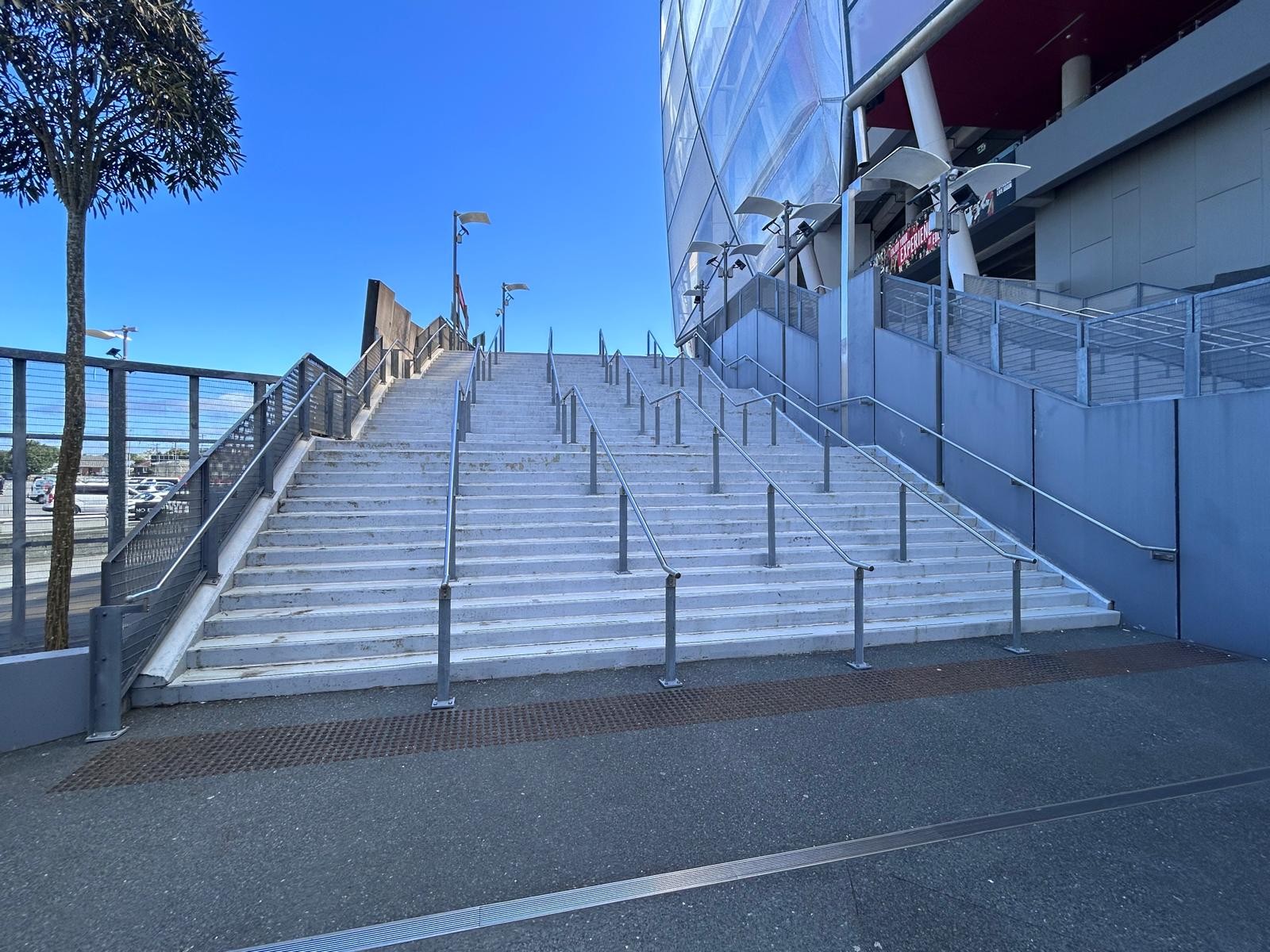Wide light-grey concrete stairs with multiple stainless steel handrails. Brown tactile paving strip at the base on a level asphalt surface leading towards a modern glass building.