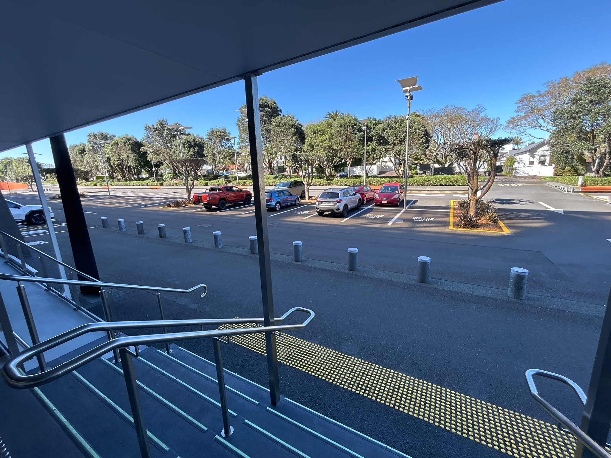 Silver metal handrails line stairs leading to level asphalt with yellow tactile ground surface indicators. Concrete bollards separate the pathway from a car park with marked bays.
