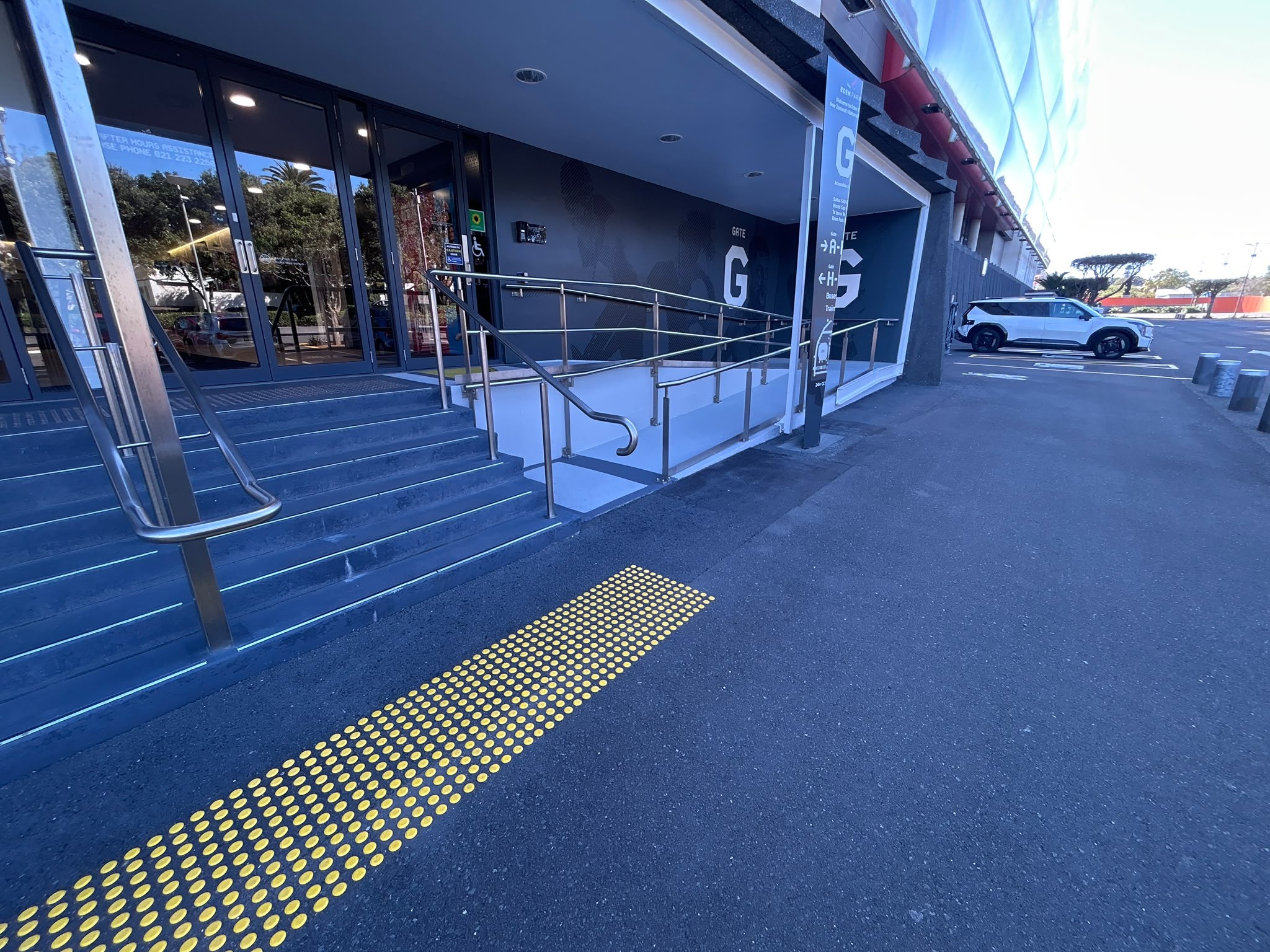 Yellow tactile paving on an asphalt path leads to a concrete ramp and stairs with silver handrails. Gate G features wide glass doors with accessibility signage on the walls and pillars.