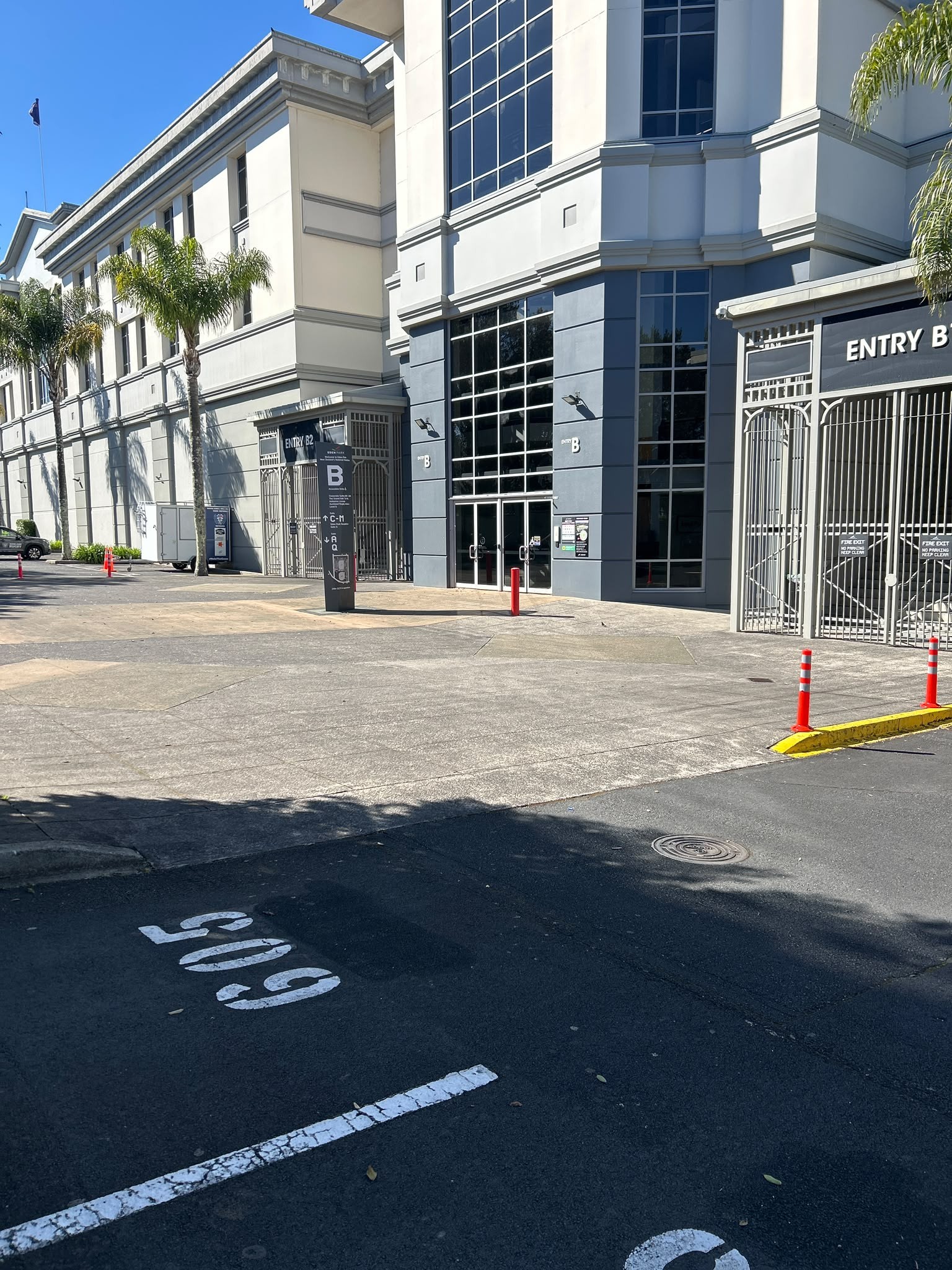 Level concrete pathway leads to glass doors at Entry B and gated Entry B2. A black pillar sign displays accessibility symbols. Orange bollards sit on a yellow kerb next to asphalt parking.