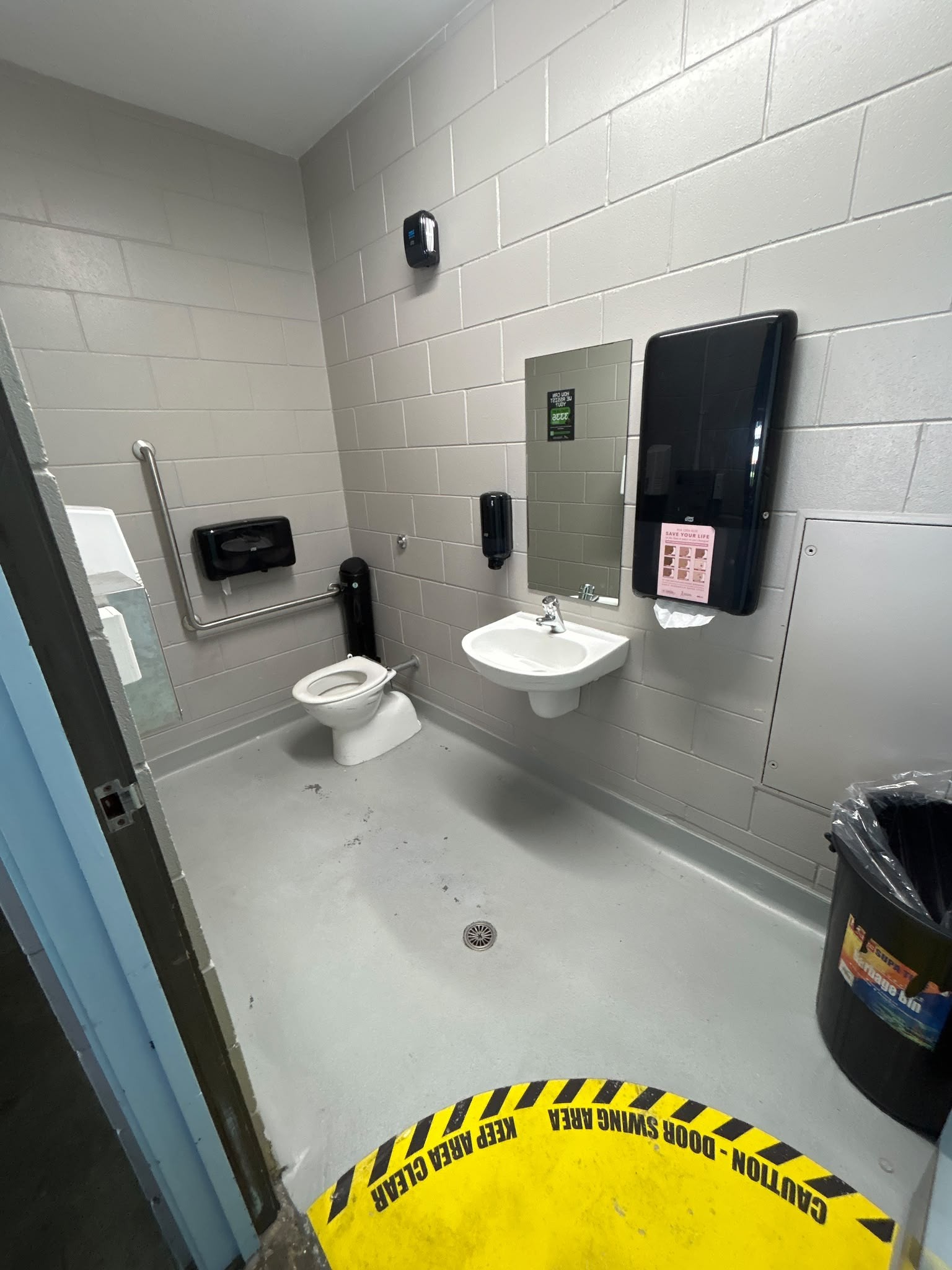 Wide accessible toilet with level grey floor. L-shaped stainless steel grab rail beside toilet. Wall-hung basin with lever tap. Yellow door-swing hazard marking on floor at entry.