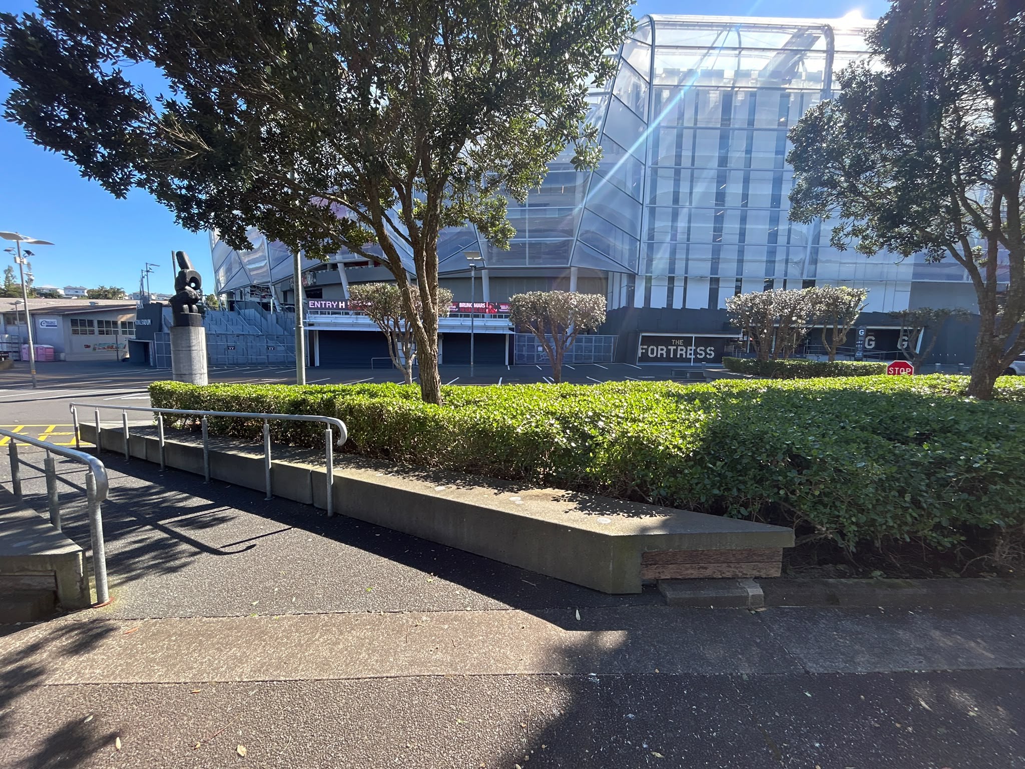 Level concrete walkway with metal handrails on a low wall. Hedges lead towards a glass-fronted stadium with 'Entry H' and 'THE FORTRESS' signs. Wide, flat ground surfaces in bright sunlight.