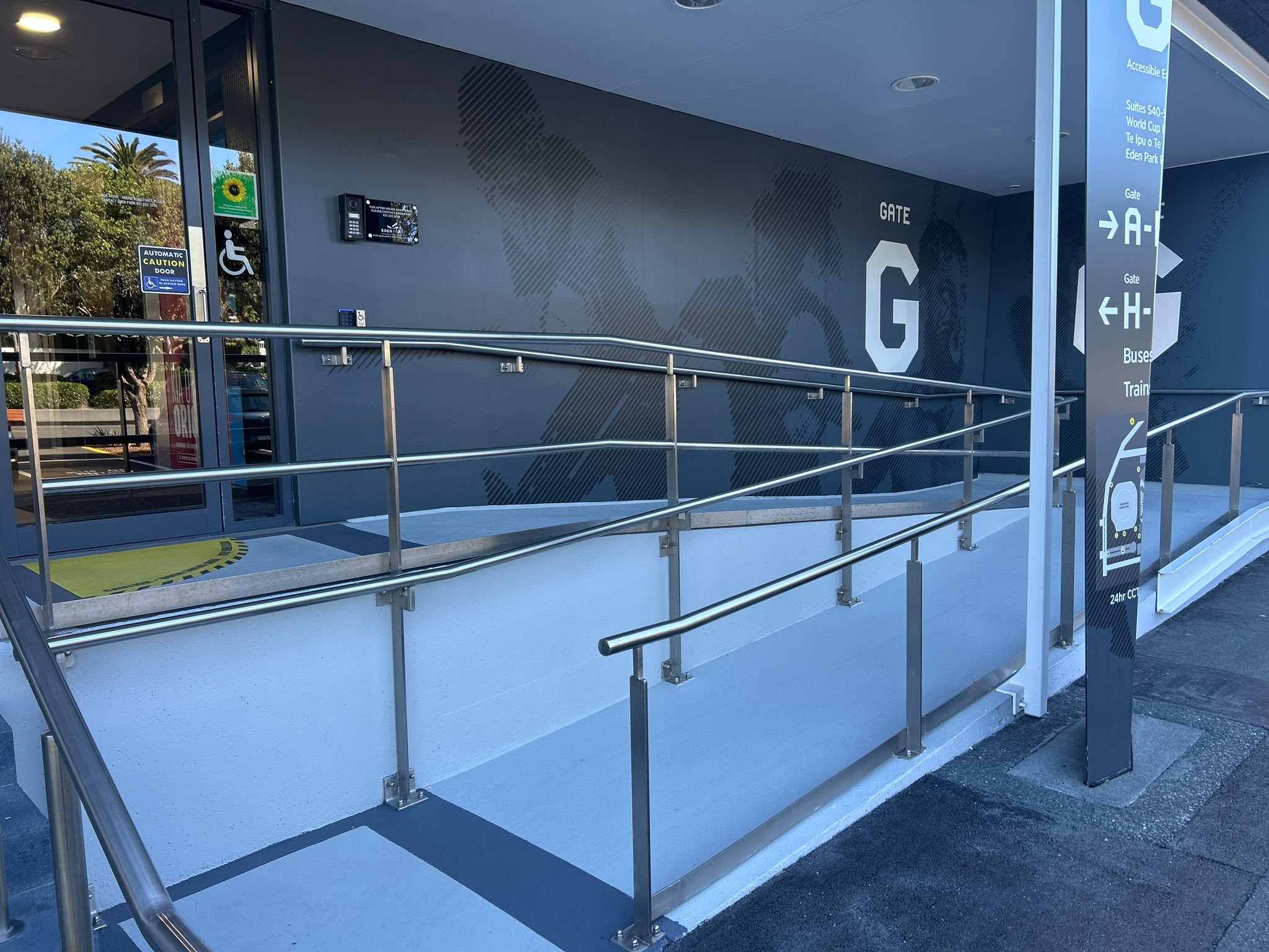 Sloping blue-grey ramp with double stainless steel handrails leads to an automatic glass door with accessibility symbols. Large white 'GATE G' sign on dark grey wall and tall directional sign.