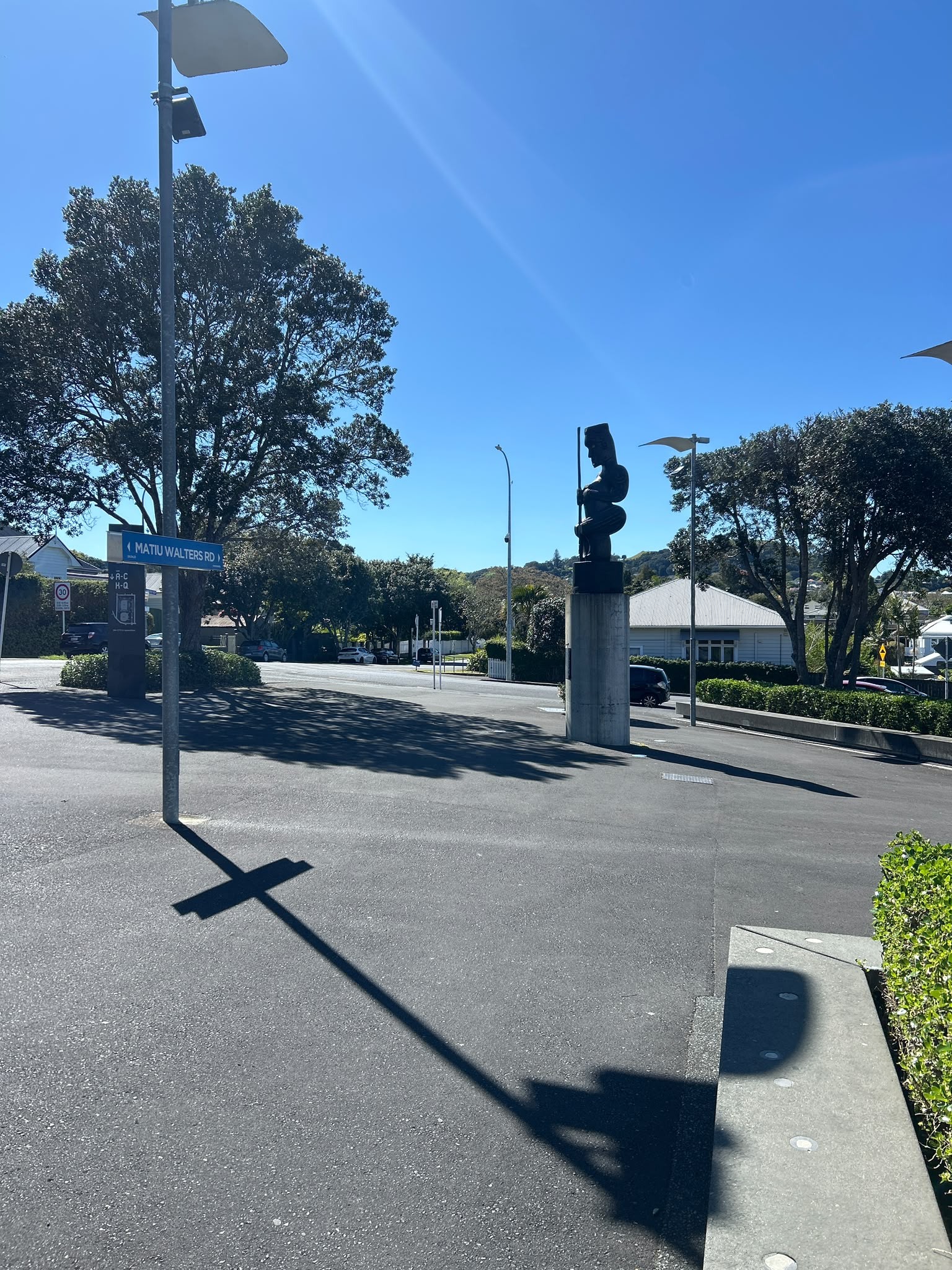 Level asphalt area with Matiu Walters Rd sign and a dark sculpture on a concrete pedestal. A low concrete ledge with inset lights and green hedges border the open space in bright sunlight.