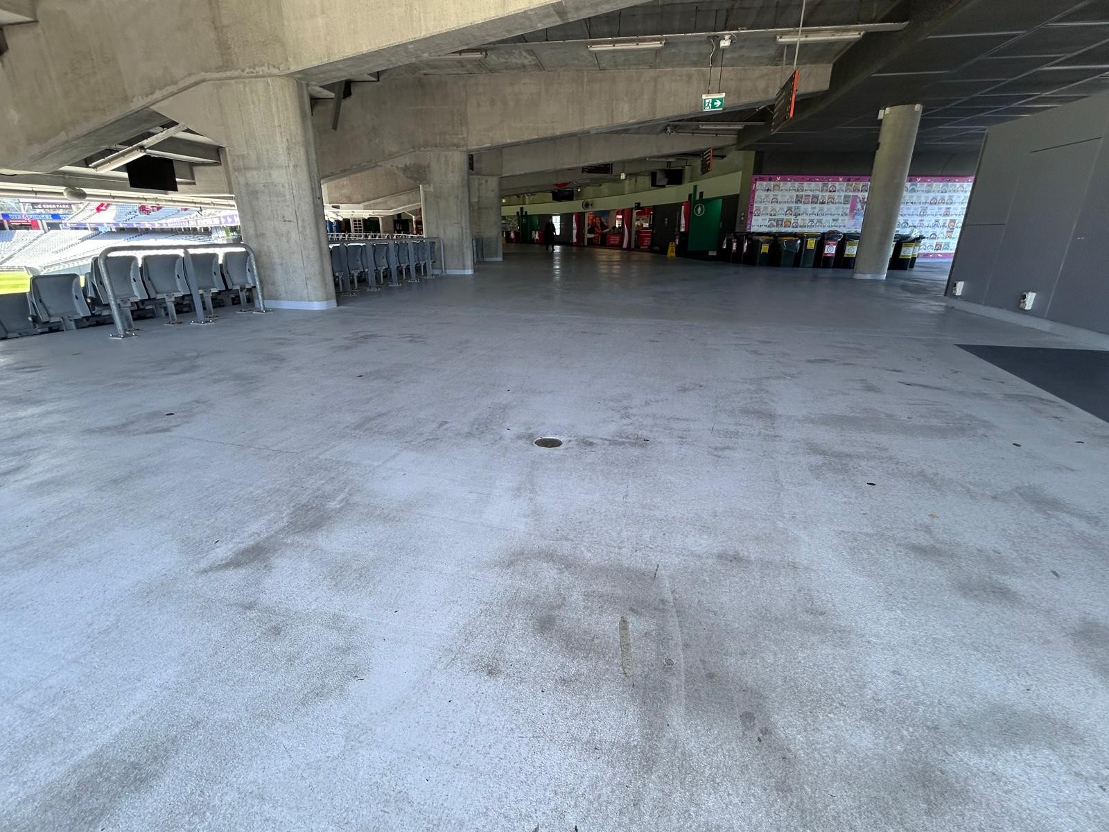 Wide, level concrete concourse with large support pillars. Rows of grey plastic seats line the left. A flat, unobstructed floor surface leads to an open area with a green exit sign.