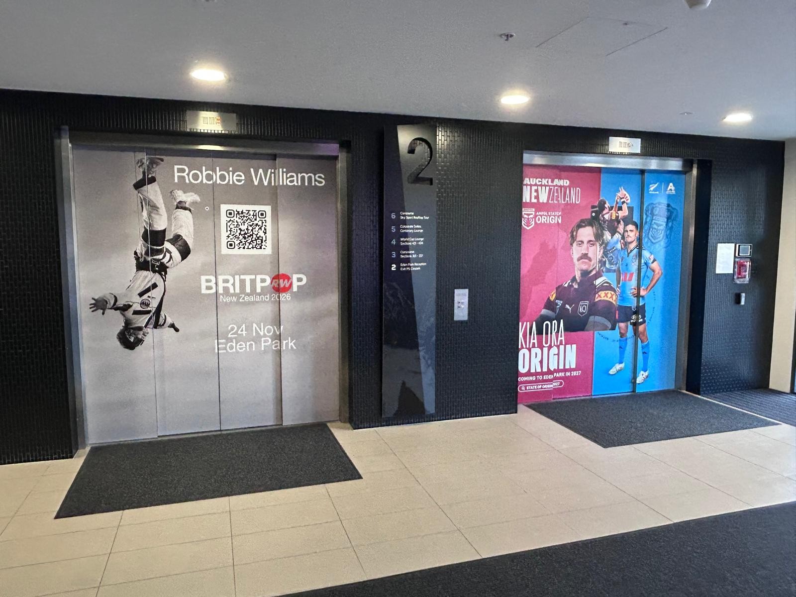 Two lift entrances on a black tiled wall with level tiled floor and dark mats. Call buttons and a floor directory are between the lifts. Wide indoor area with bright overhead lights.