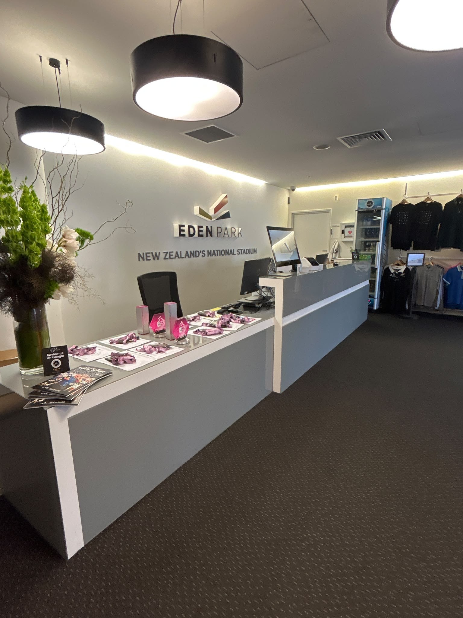 Grey reception desk with white trim on level dark grey carpet. Large 'EDEN PARK' signage on wall. Bright pendant lighting overhead. Flat, unobstructed floor path to the counter.