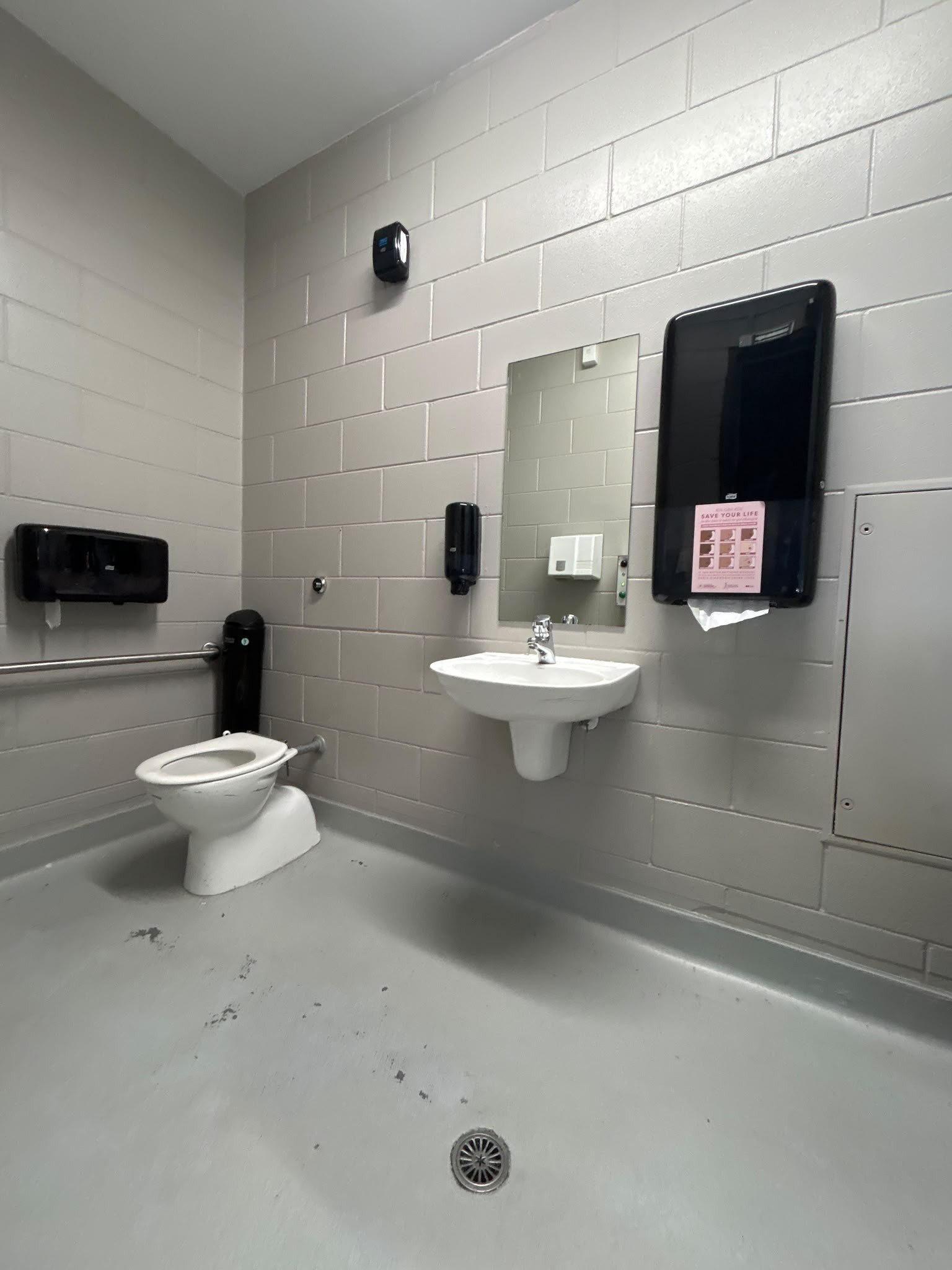 Accessible restroom with grey block walls and level floor. White toilet has a horizontal grab rail. Wall-hung basin with chrome tap and mirror. Black dispensers and central floor drain.