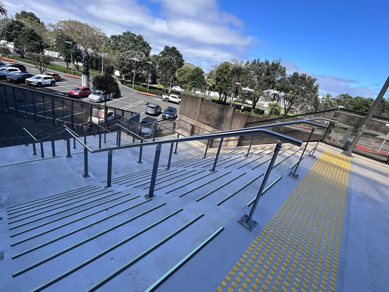 Wide concrete outdoor stairs with dark anti-slip strips on step edges. Metal handrails on sides and centre. Yellow tactile ground surface indicators at the top right.