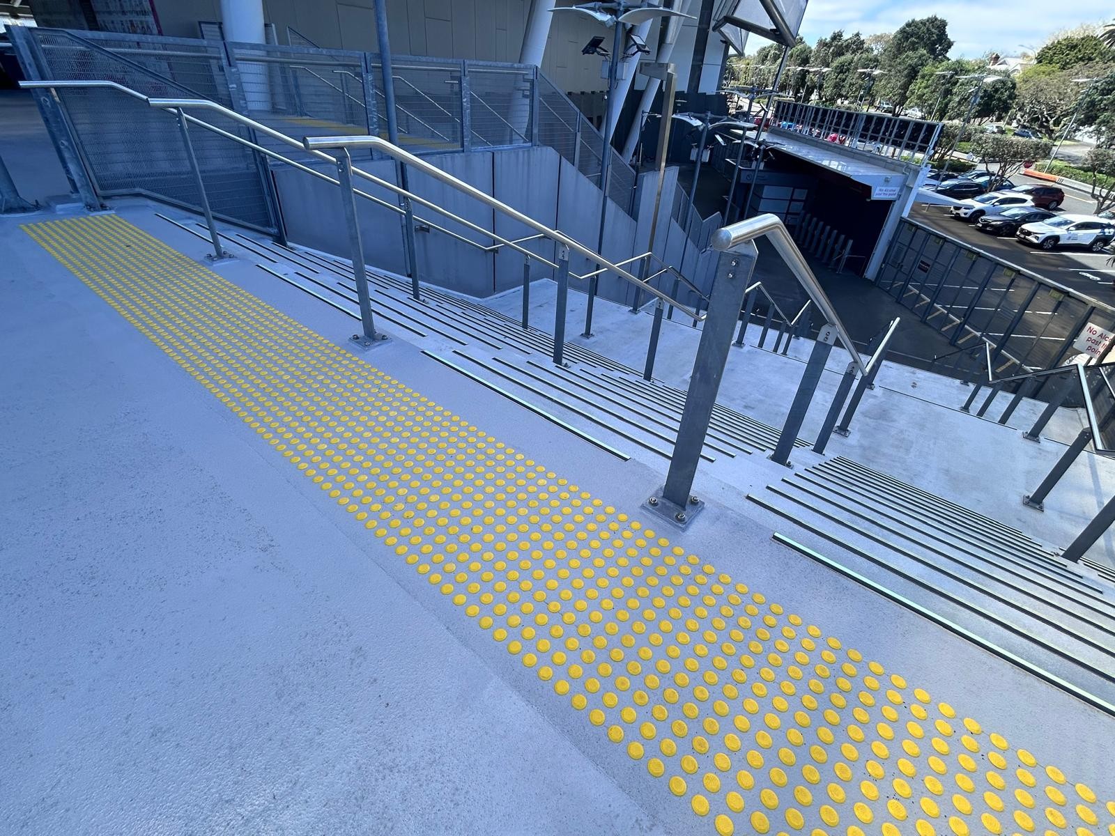 Wide light grey concrete stairs with yellow tactile paving at the top landing. Multiple silver metal handrails divide the stairs. Each step has a black high-contrast strip on the leading edge.