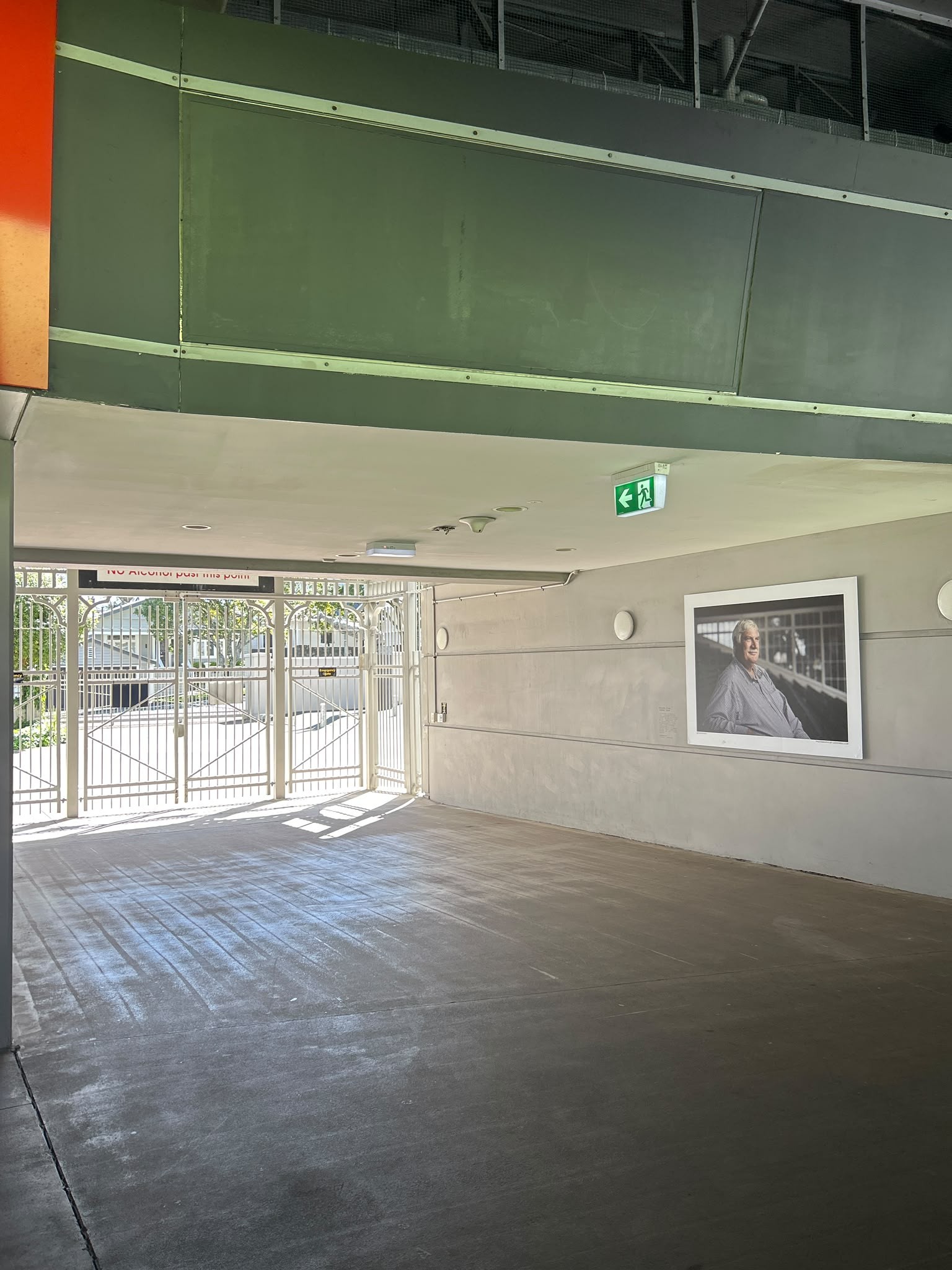 Wide, level concrete passage leading to a white metal gate. Clear, unobstructed pathway with smooth flooring, light grey walls, a large portrait, and a green exit sign pointing left.