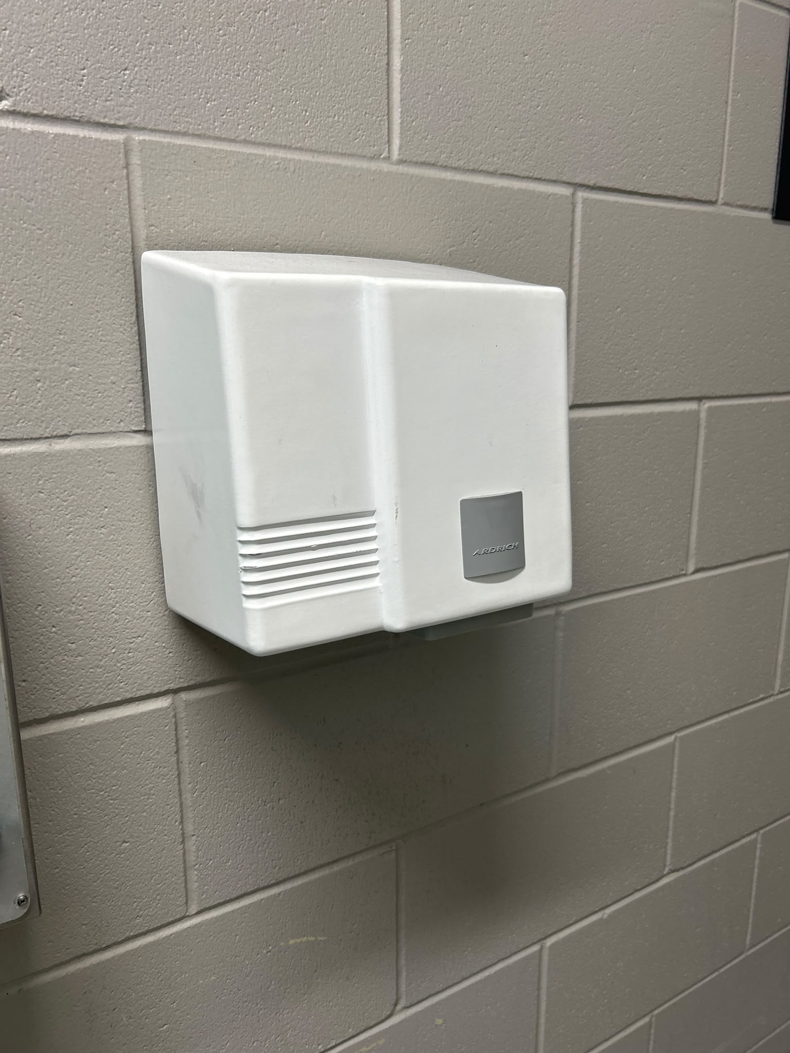 White wall-mounted hand dryer on a grey concrete block wall. Features horizontal vents on the left and a silver label. No visible buttons are present on the unit's smooth surface.