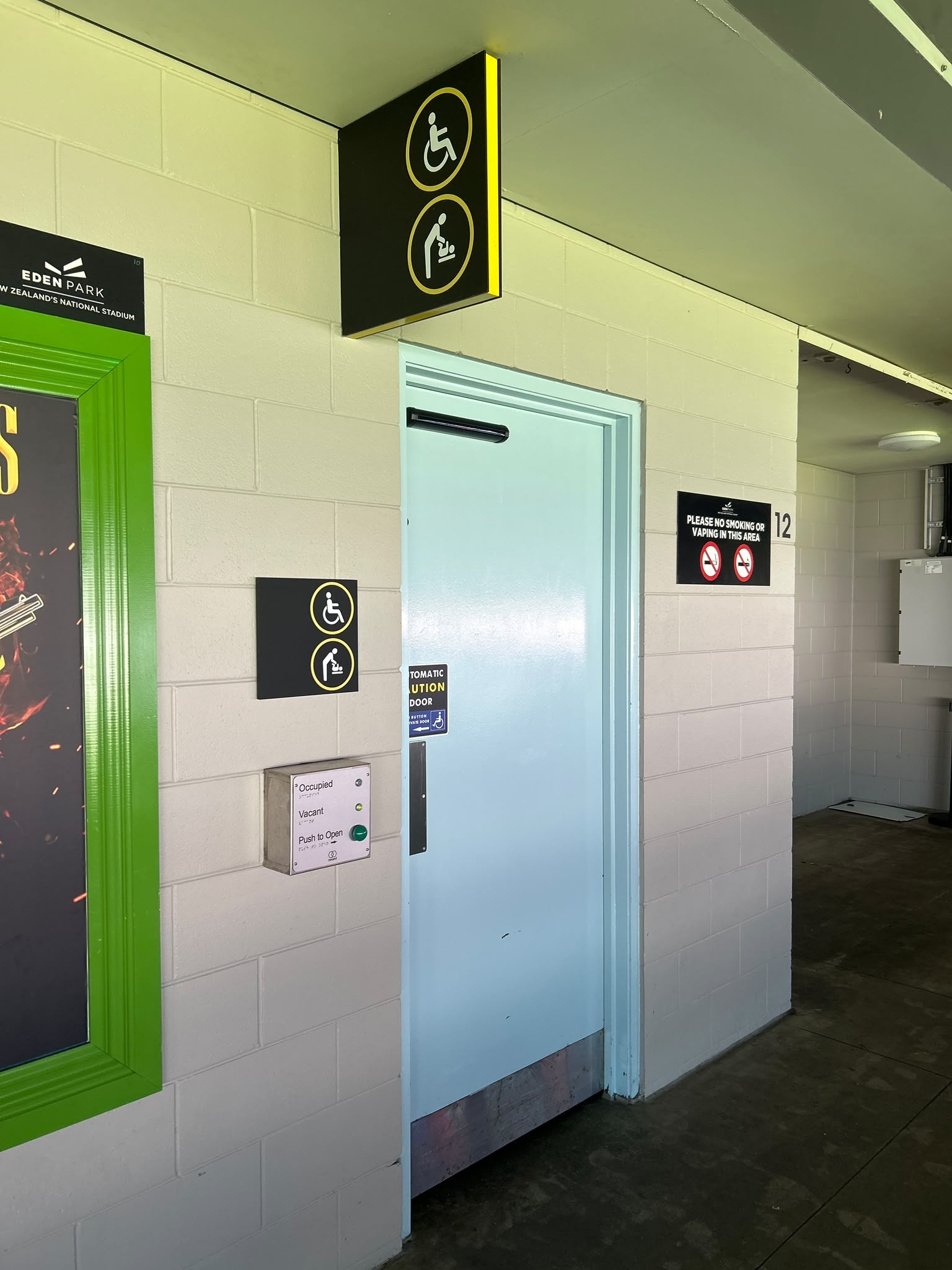 Accessible toilet with light blue automatic door and level floor. Wall panel features a green push-to-open button, status lights, and Braille. Signage shows wheelchair and baby-change symbols.