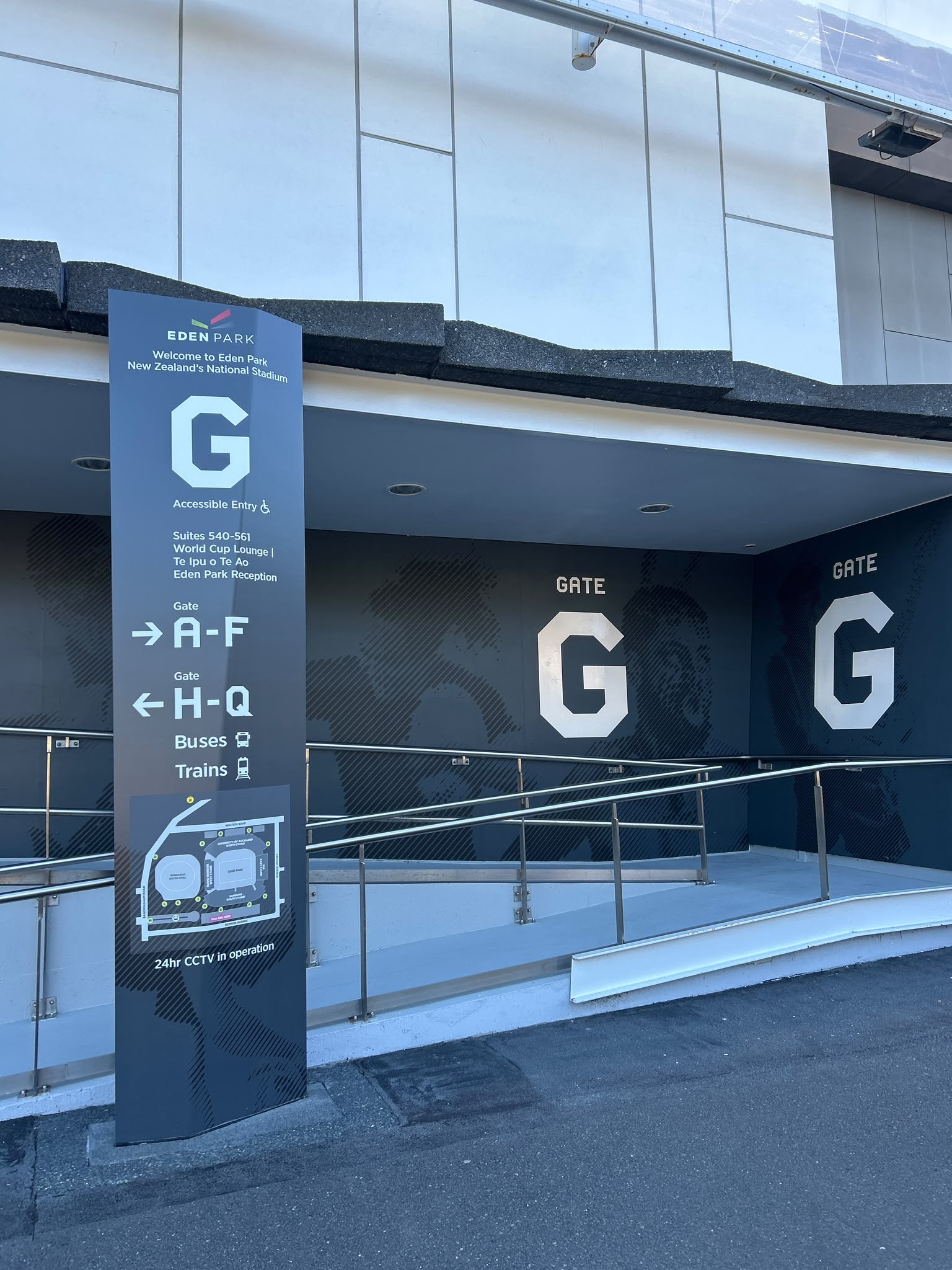Tall grey totem sign marks Gate G as an accessible entry. A concrete ramp with metal handrails leads into the building under a lit overhang. Level asphalt and concrete ground surfaces.